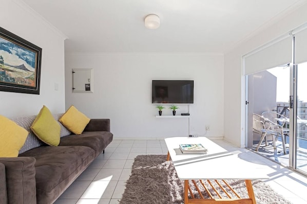 A modern living room with a sofa, a coffee table, a flat-screen TV, and a balcony with outdoor seating.