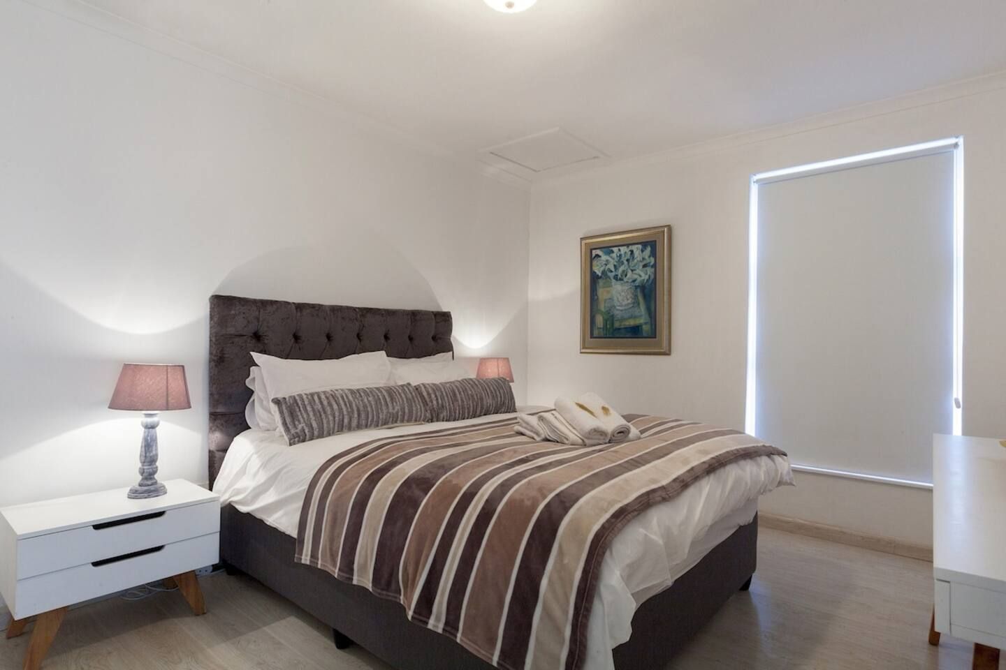 Un dormitorio con cama, mesitas de noche, lámpara, un cuadro y una puerta.