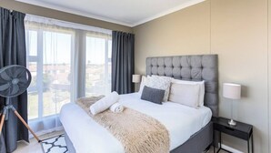 Una camera d'albergo con un grande letto, una ventola da terra, vista dalla finestra e un comodino con una lampada.