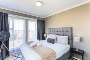 Una habitación de hotel con una cama grande, un ventilador de pie, vistas por la ventana y una mesita de noche con lámpara.