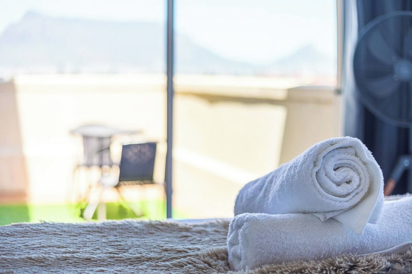 A neatly rolled white towel on a textured rug, a small table with chairs, and a fan in the background.