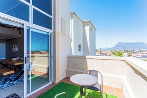 Un balcon avec une table et des chaises, une porte-fenêtre coulissante et une vue sur un paysage urbain avec des montagnes à l’horizon.