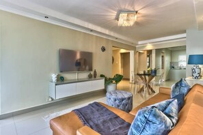 A modern living room with a flat-screen TV, a sofa, ottoman, and a dining area with a glass table.