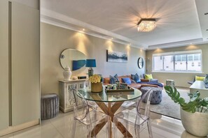Una sala moderna con una mesa de centro de vidrio, sillas transparentes, un sofá con cojines, un espejo decorativo y una araña.