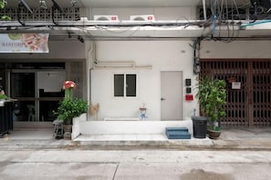 A building with a white facade, a small entrance, and a door with a window. There are potted plants on the sidewalk. - Bangkok Group Haven - 6BR Bangchak BTS 12 sleeps (Bangkok)