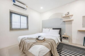 A bedroom with a bed, a bedside table, a lamp, a window with blinds, and a wall-mounted air conditioner. - Bangkok Group Haven - 6BR Bangchak BTS 12 sleeps (Bangkok)