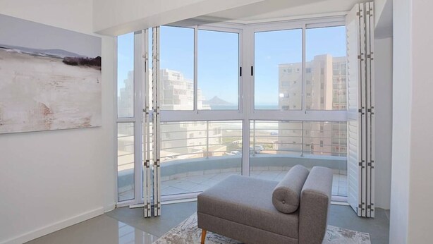 A modern living room with a sofa, a large window with a view of buildings, and a painting on the wall. - 3br Beach Stay | Views & backup power by ctha (Cape Town)