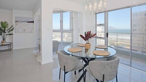 A modern dining area with a glass table, four chairs, and a view of the city through large windows. - 3br Beach Stay | Views & backup power by ctha (Cape Town)