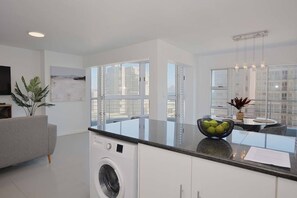 A modern kitchen with a built-in washer, large windows, and a dining area with a bowl of fruit.