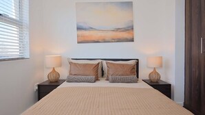 A bedroom with a large bed, two bedside tables with lamps, a painting above the bed, and a window with blinds. - 3br Beach Stay | Views & backup power by ctha (Cape Town)