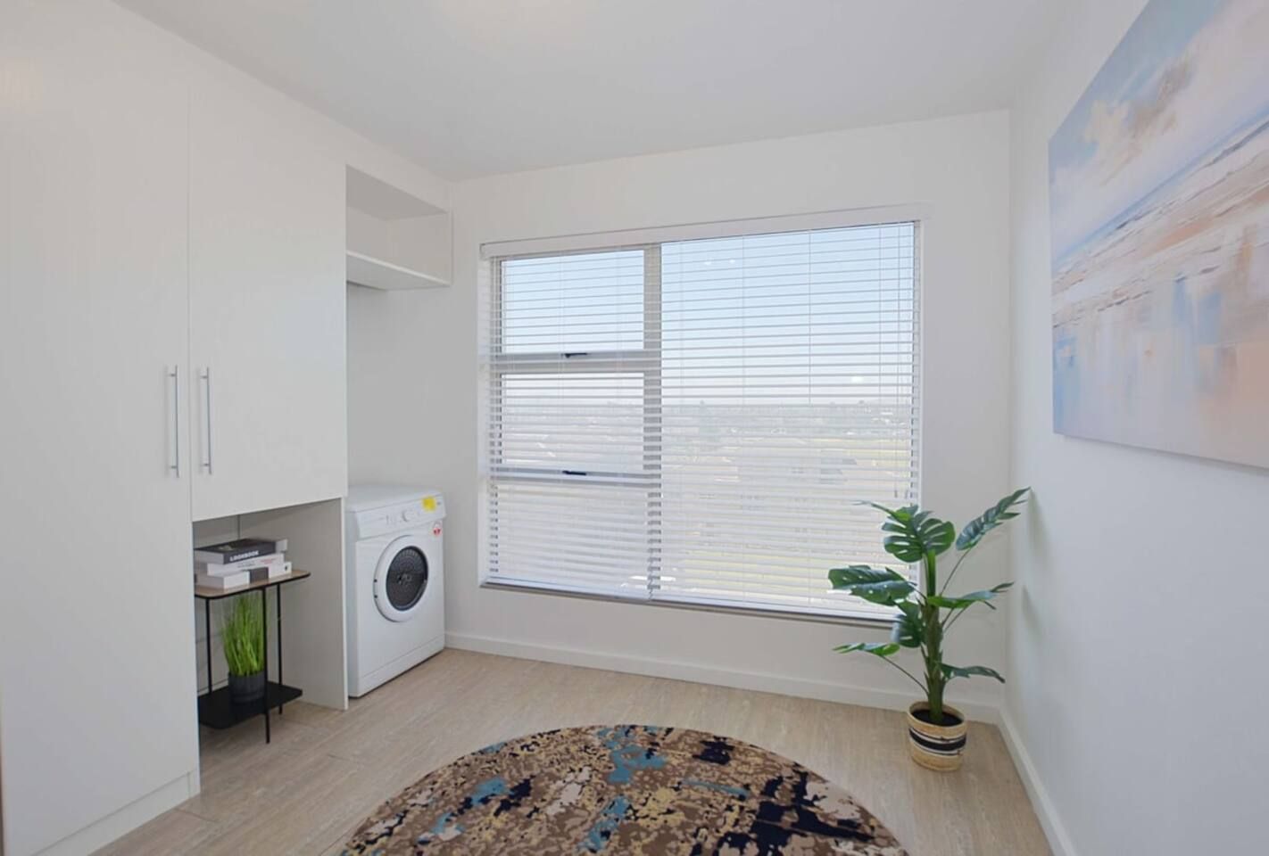 A laundry room with a washing machine, a window with blinds, a potted plant, and a painting on the wall.