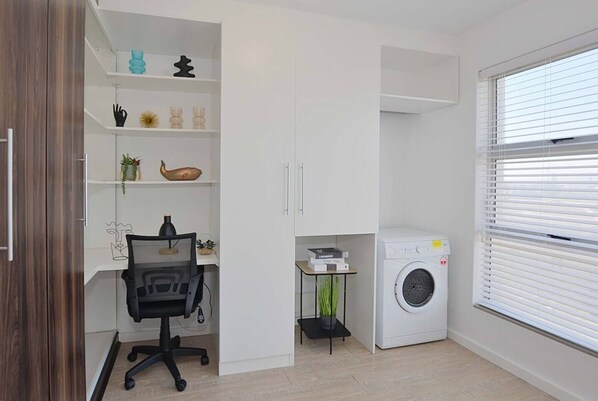 A compact living space with a desk, chair, shelving with decorative items, a washing machine, and a window with blinds.