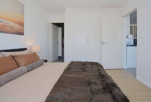 A bedroom with a bed, a fur throw, a painting, and a door leading to a kitchen.