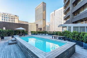 Una piscina sul tetto con un pontile in legno, circondata dal verde e con moderni edifici sullo sfondo.