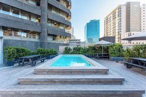 A rooftop pool area with steps leading to the pool, surrounded by greenery and modern buildings in the background.
