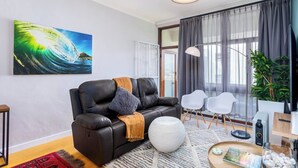 A modern living room with a leather sofa, a round white ottoman, a large painting, and a view through a glass door. - Burmeister Circle by Ctha (Milnerton)