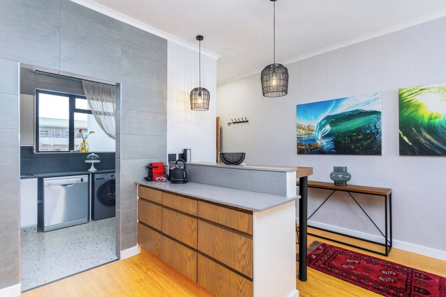 A modern kitchen with a built-in dishwasher, a coffee machine, and a kettle on the counter. There are two pendant lights above the counter and a red rug on the floor.