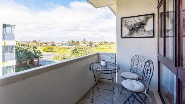 A balcony with a view of a residential area, a small table with a chair, and a framed artwork on the wall. - Burmeister Circle by Ctha (Milnerton)