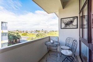 A balcony with a view of a residential area, a small table with a chair, and a framed artwork on the wall.