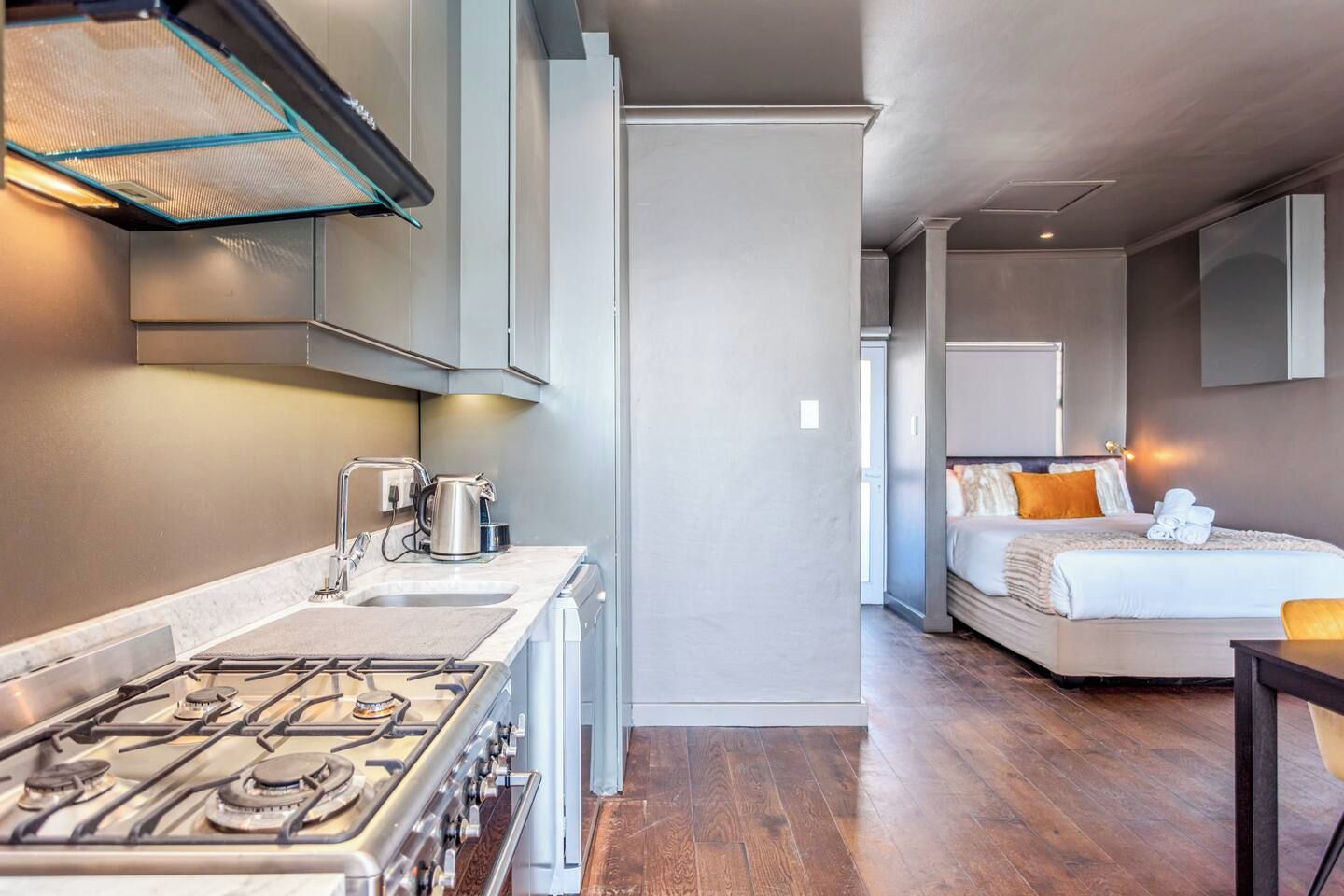 A modern kitchen with a gas stove and a coffee maker, leading to a bedroom with a bed and a nightstand.