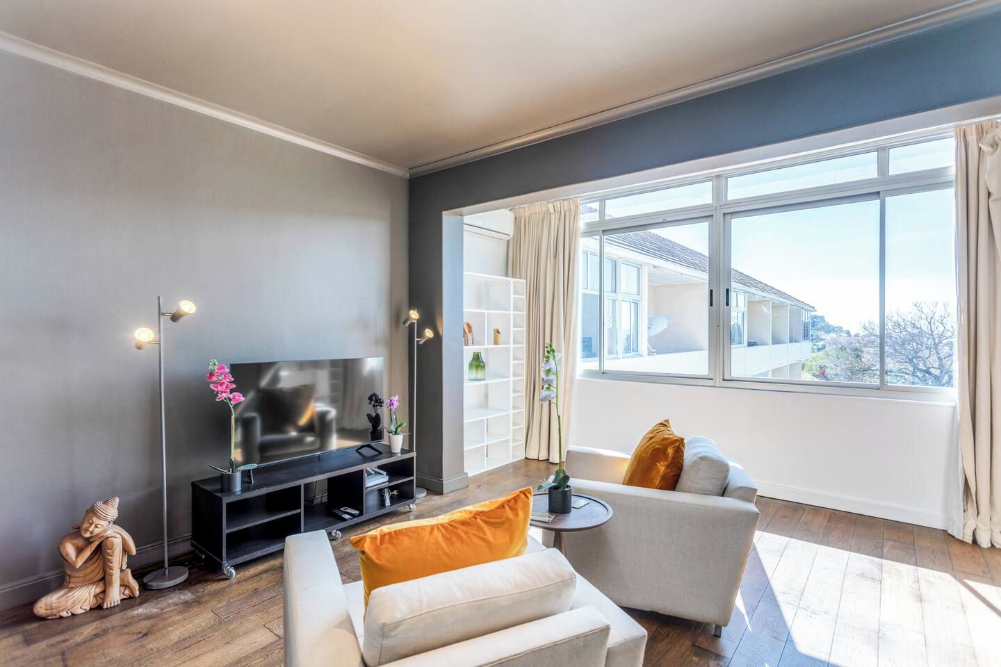 Ein modernes Wohnzimmer mit einem Flachbildfernseher, einem Sofa, einem Couchtisch und einem großen Fenster mit Blick auf ein Gebäude.