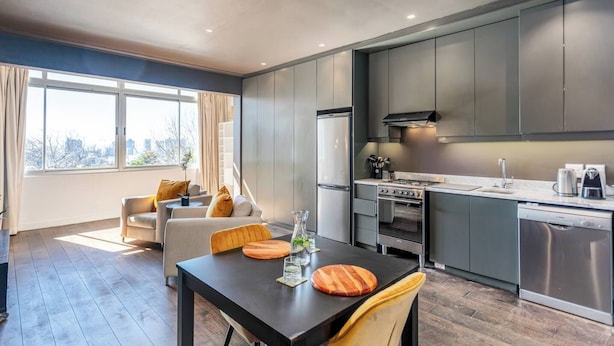 A modern kitchen with built-in appliances, a dining area with a table and chairs, and a view of the city through large windows. - Granada Court 309 by Ctha (Cape Town)