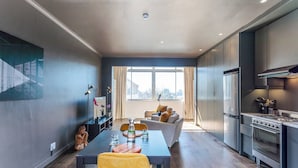 A modern kitchen with stainless steel appliances, a dining area with a table and chairs, and a living space with a sofa and a large window. - Granada Court 309 by Ctha (Cape Town)