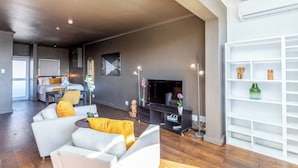 A modern living room with a sofa, a coffee table, and a TV. There is a bookshelf and a dining area in the background. - Granada Court 309 by Ctha (Cape Town)