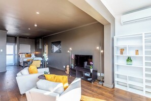 Un soggiorno moderno con un divano, un tavolino da caffè e una TV. Si nota una libreria e un'area pranzo sullo sfondo.