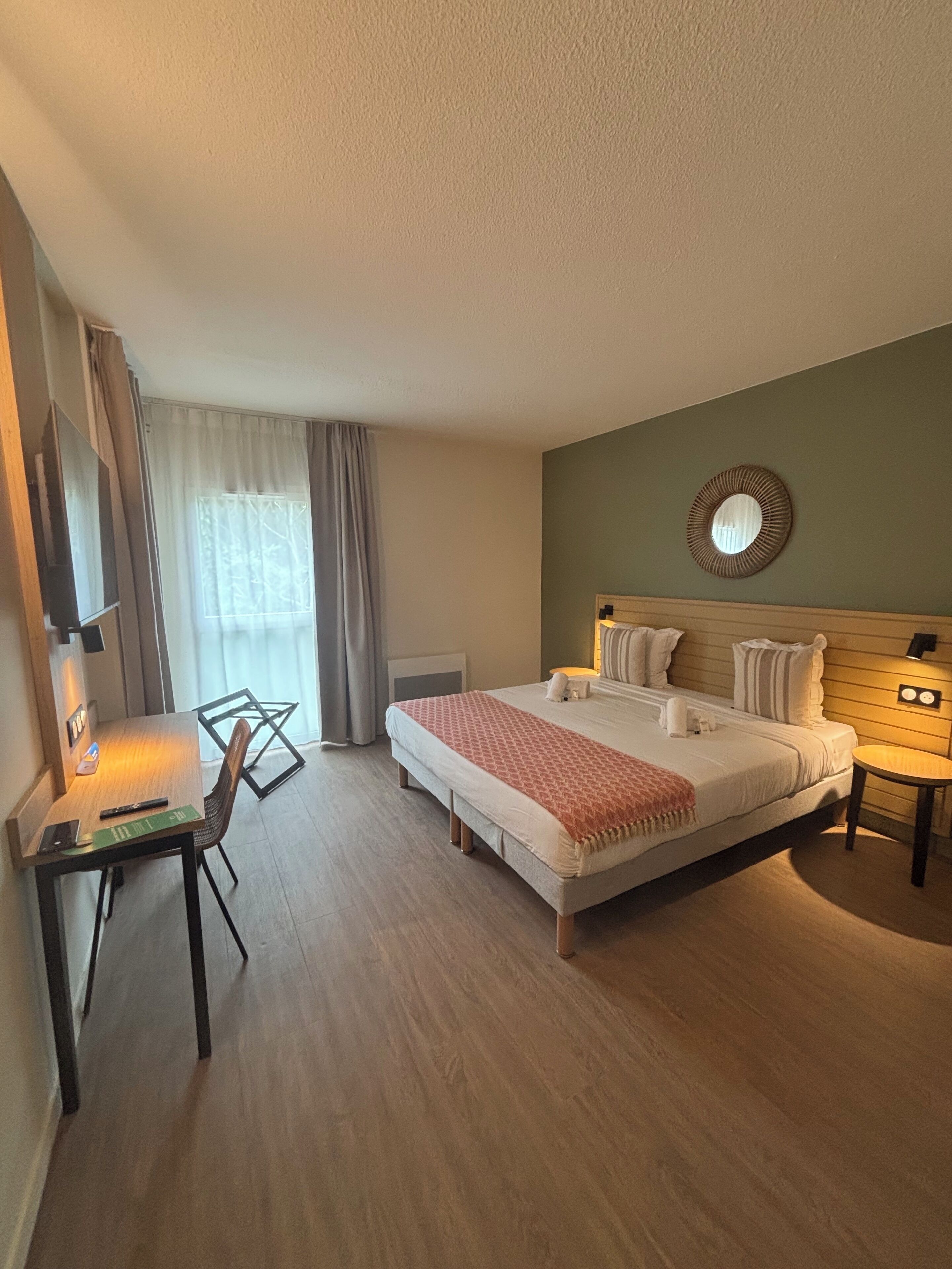 A hotel room with a bed, bedside tables, a desk with a chair, a TV, and a window with curtains.