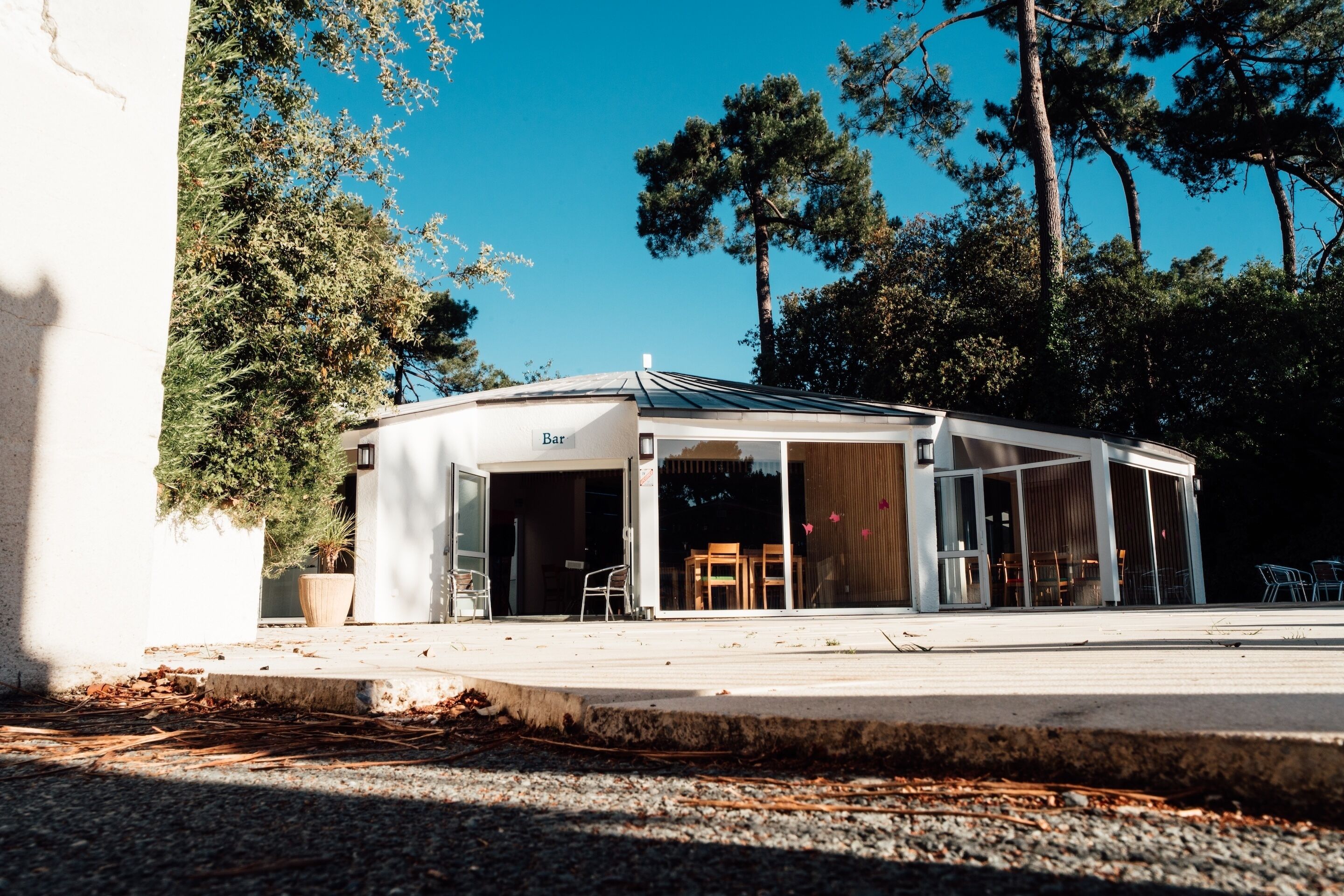 Un bar extérieur moderne, doté d’une structure en verre, entouré d’arbres et bordé d’un chemin de gravier.