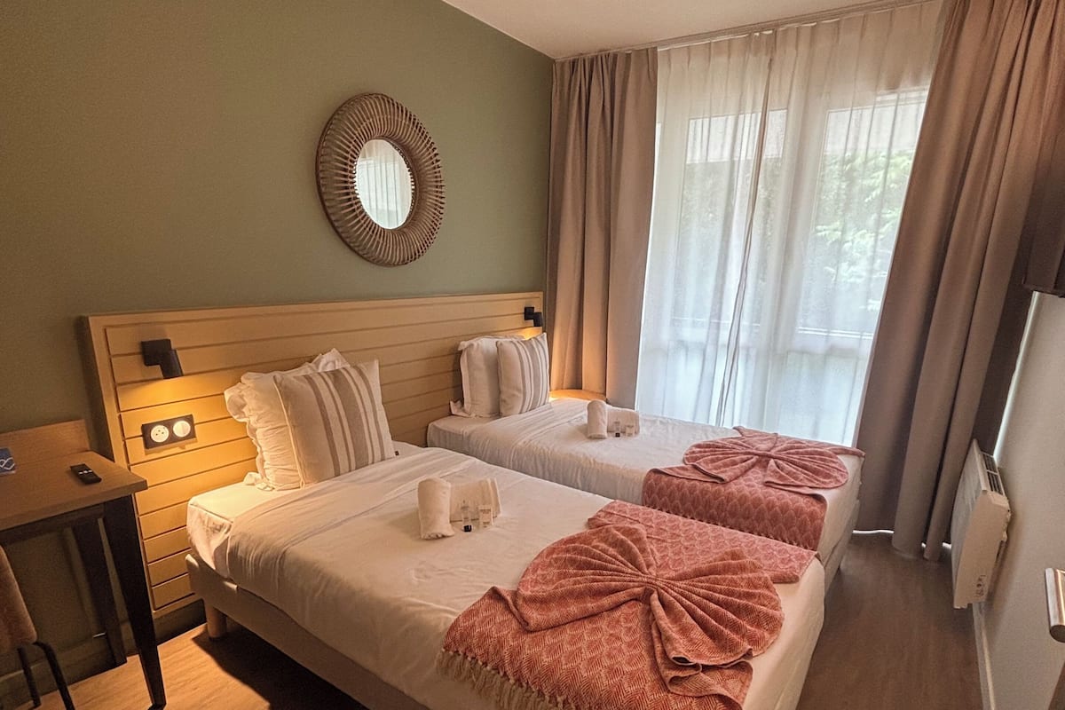 A hotel room with two beds, a wooden headboard, a small table with a chair, a mirror, and a window with curtains.