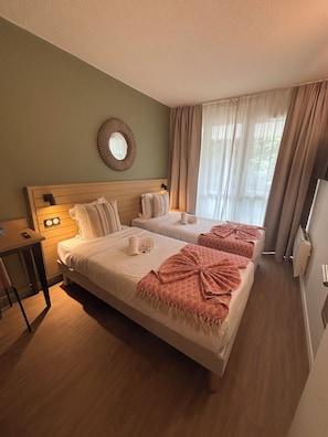 Ein Hotelzimmer mit zwei Betten, einem Holzkopfende, einem kleinen Tisch mit Stuhl, einem Spiegel und einem Fenster mit Vorhängen.