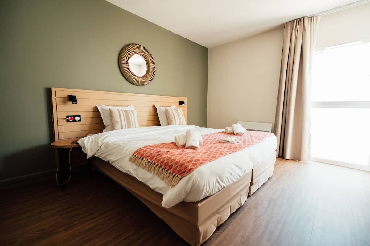 A neatly made bed with a headboard, a bedside table, a wall-mounted mirror, and a window with curtains.