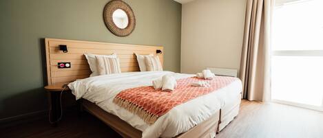 A neatly made bed with a headboard, a bedside table, a wall-mounted mirror, and a window with curtains.