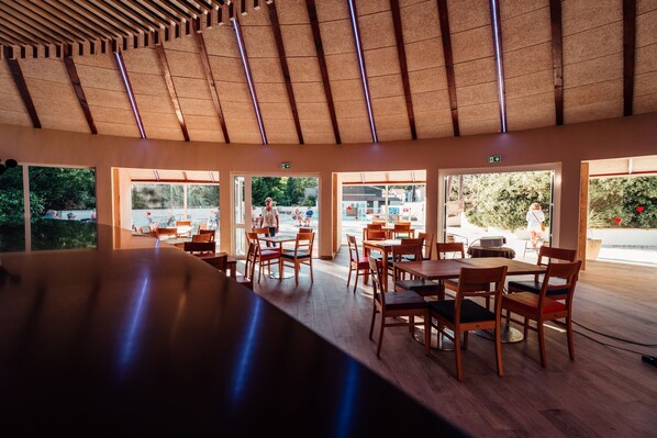 An indoor dining area with wooden tables and chairs, large windows, and a high ceiling with wooden beams. - Club Vacances Bleues Le Grand Air (Saint-Trojan-les-Bains)