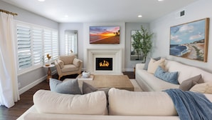 A living room with a fireplace, a sofa, a coffee table, and a painting of a beach scene. - Modern SoCal Escape with Outdoor Grill & Lush Lounge Areas | Casa Cordero (Mission Viejo)