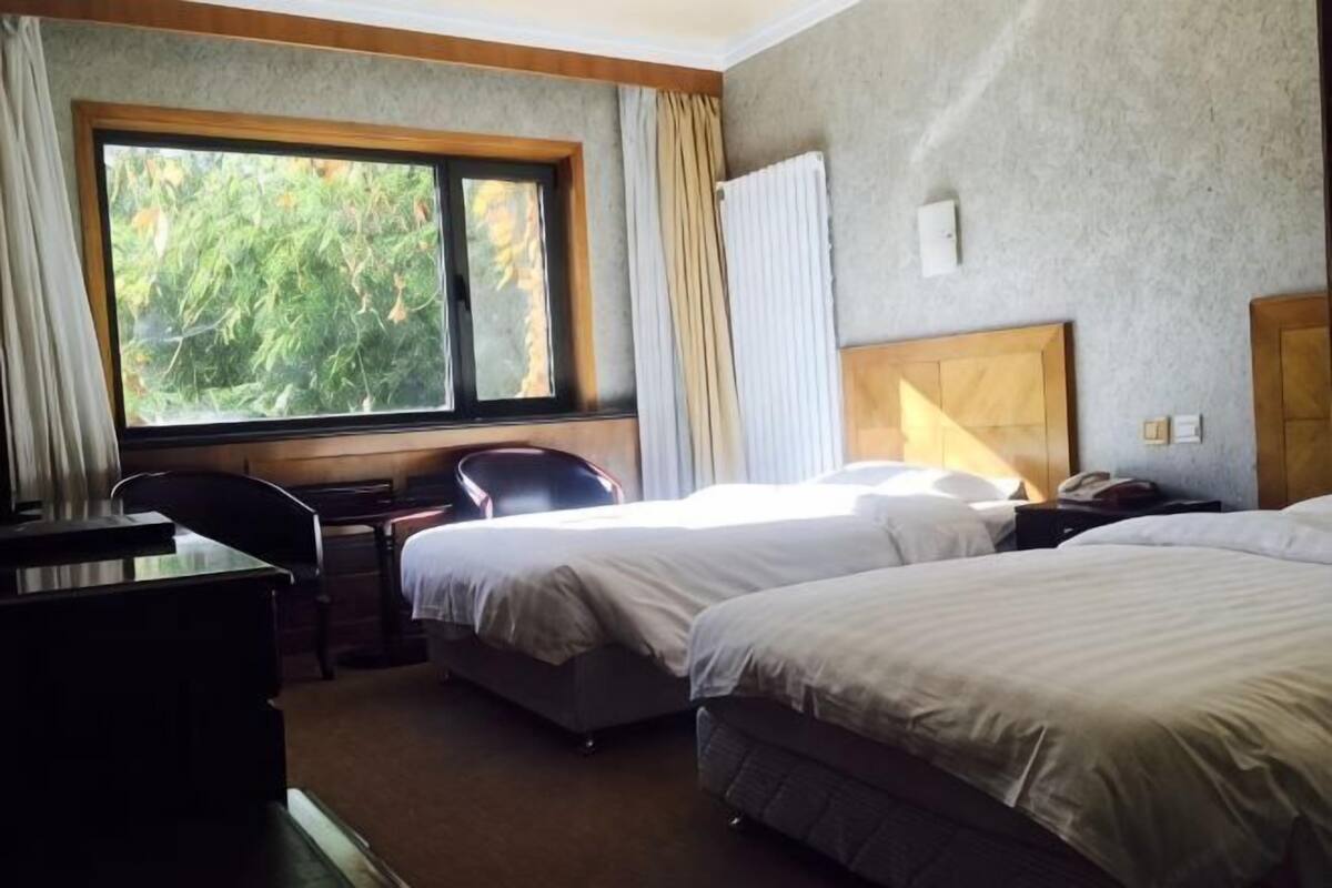 Une chambre d’hôtel avec deux lits, un bureau et une chaise. Une fenêtre offre une vue sur les arbres.