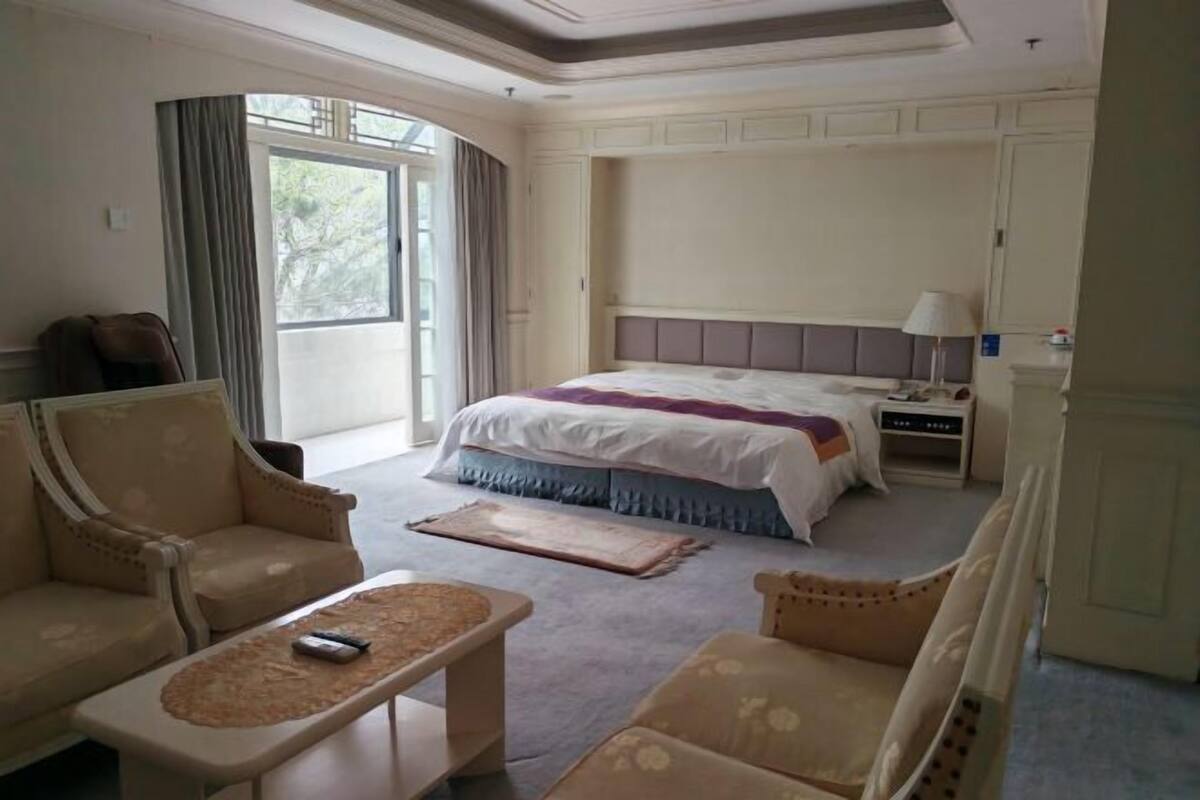 Une chambre d’hôtel dotée d’un grand lit, d’un coin salon avec un canapé et des fauteuils, d’une petite table et d’une fenêtre avec des rideaux.