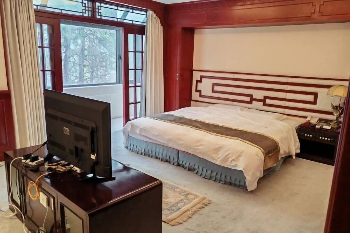 Une chambre à coucher avec un lit, un téléviseur posé sur un meuble en bois et une fenêtre avec des rideaux.
