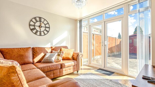 A living room with a brown leather sofa, a wall clock, and a large sliding glass door leading to an outdoor area. - Spacious & Modern 4-Bed Home – Sleeps 7 (Newquay)