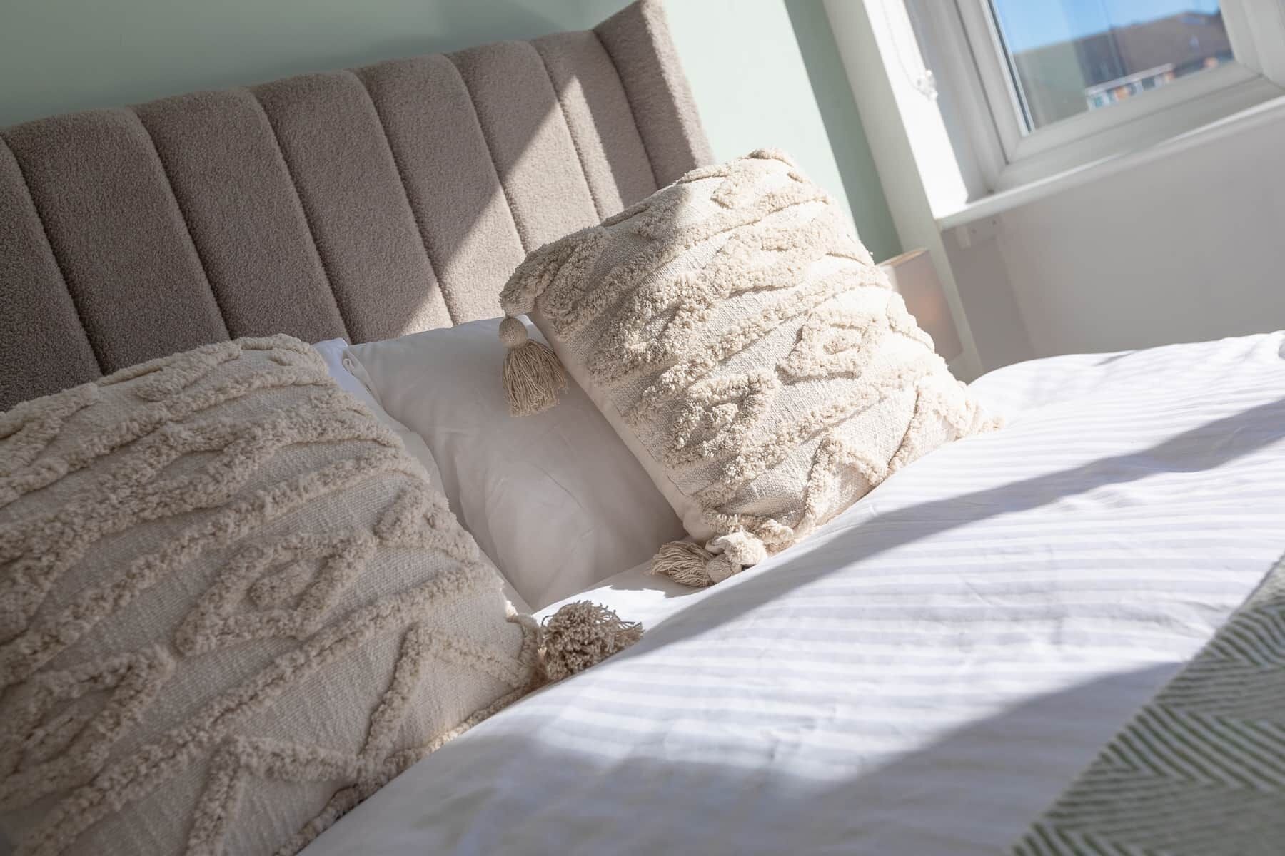 Un lit avec une couverture et des oreillers de couleur beige à texture, une tête de lit et une fenêtre donnant sur des immeubles.