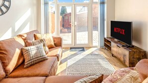 A living room with a brown leather sofa, a flat-screen TV displaying 'NETFLIX', and a sliding glass door leading to an outdoor area. - Spacious & Modern 4-Bed Home – Sleeps 7 (Newquay)