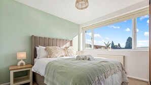 A bedroom with a large bed, a bedside table with a lamp, a window with a view of the sky and trees, and a radiator under the window. - Spacious & Modern 4-Bed Home – Sleeps 7 (Newquay)