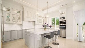 A modern kitchen with white cabinetry, a large island with bar stools, pendant lighting, and a view of greenery outside.