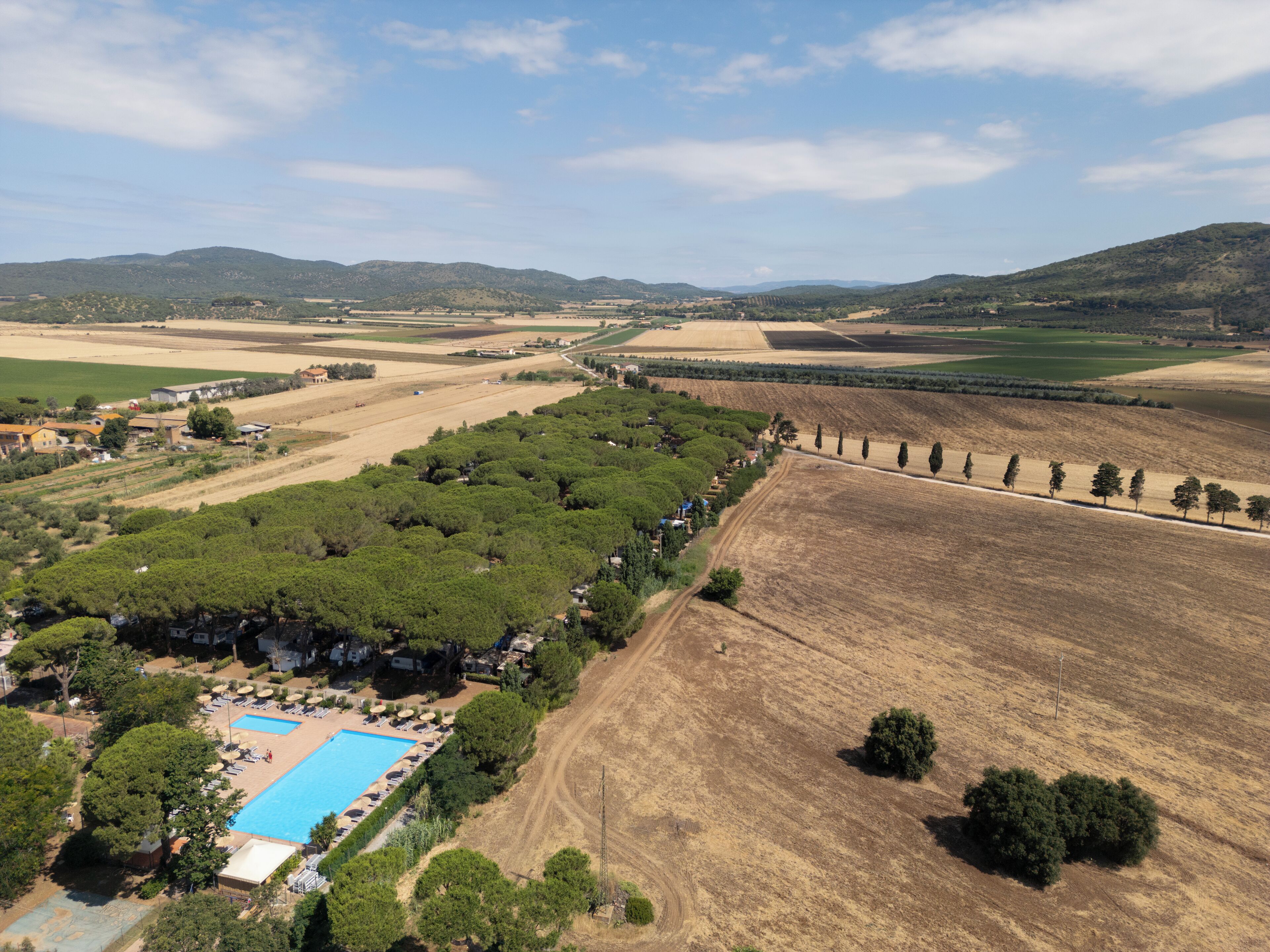 Vue aérienne d’un complexe hôtelier doté d’une piscine, entouré d’une végétation luxuriante et de champs.