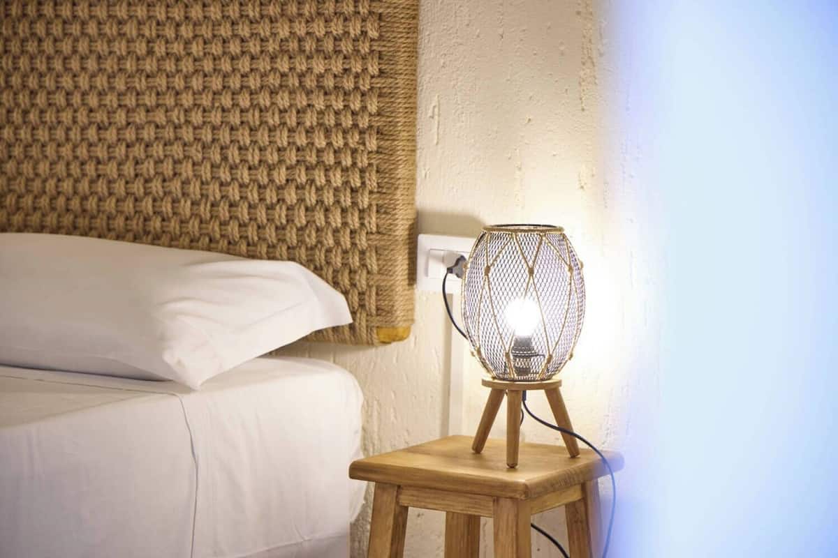 Ein Bett mit weißen Leinen und einem gewebten Kopfteil, ein kleiner Nachttisch mit einer Drahtlampenleuchte und eine Steckdose.