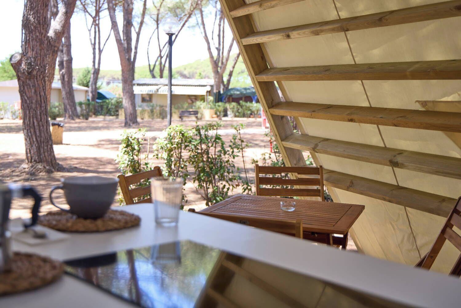 A wooden table with a glass and a cup, a chair, and a view of trees and a building in the background.
