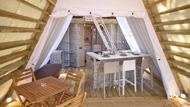 Un interior acogedor de una estructura en forma de cúpula de madera, con comedor, pequeña cocina y una escalera que lleva a un nivel superior.