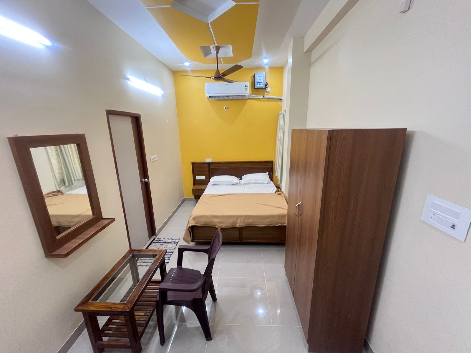 A compact hotel room with a bed, a desk with a chair, a wardrobe, and a ceiling fan.